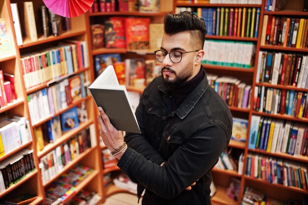 L'uomo alto e intelligente dello studente arabo indossa una giacca di jeans nera e occhiali da vista in biblioteca con un libro a portata di mano