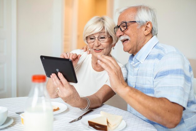 L'uomo anziano e sua moglie stanno usando una tavoletta digitale e comunicano con i loro figli La coppia senior felice sta facendo colazione e ha una conversazione online con la propria famiglia e inviando saluti