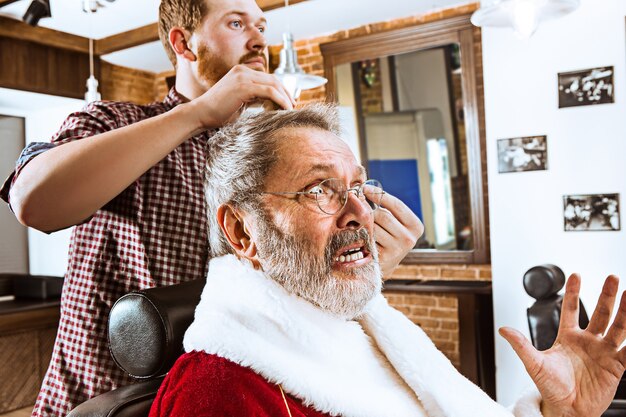 L'uomo anziano in costume di Babbo Natale che rade il suo maestro personale al negozio di barbiere prima di Natale