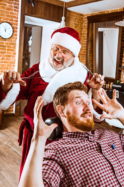 L'uomo anziano in costume di Babbo Natale lavora come maestro personale con le forbici al negozio di barbiere prima di Natale