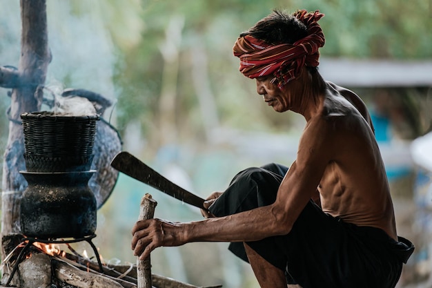L'uomo anziano senza maglietta e il perizoma con turbante usa il legno tagliato con il coltello per fare legna da ardere riso appiccicoso cotto a vapore, vita semplice della gente rurale, spazio per la copia, scena rurale della campagna in Thailandia