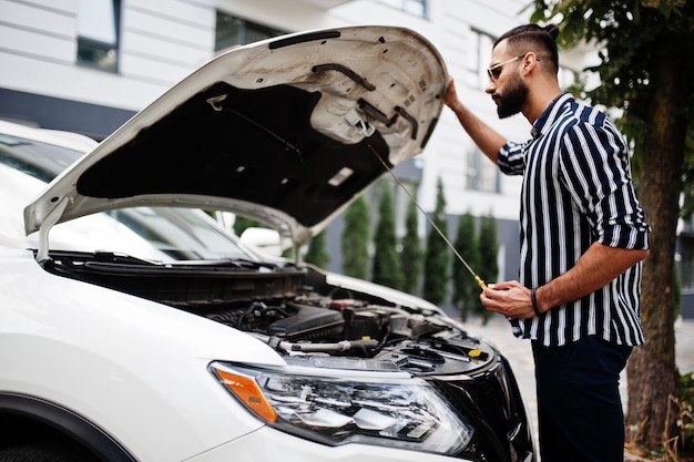 L'uomo arabo di successo indossa una camicia a righe e occhiali da sole posa vicino al suo motore di controllo dell'auto suv bianco con cappuccio aperto