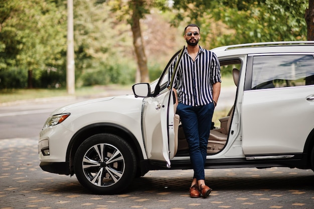 L'uomo arabo di successo indossa una camicia a righe e occhiali da sole posa vicino alla sua auto bianca suv Eleganti uomini arabi in trasporto