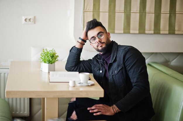 L'uomo arabo indossa una giacca di jeans nera e occhiali da vista seduto al bar, legge un libro e beve un caffè Ragazzo modello arabo elegante e alla moda