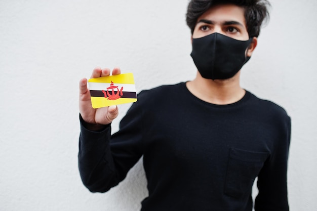 L'uomo asiatico indossa tutto nero con maschera facciale tiene in mano la bandiera del Brunei isolata su sfondo bianco Concetto di paese di coronavirus
