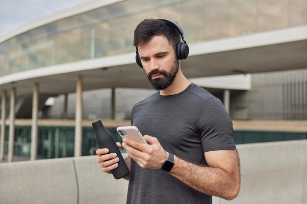 L'uomo atletico muscoloso guarda lo schermo dello smartphone scarica i brani nella playlist per l'allenamento tiene una bottiglia con acqua fresca indossa una maglietta casual ha pose di allenamento regolari all'aperto. Uno stile di vita sano