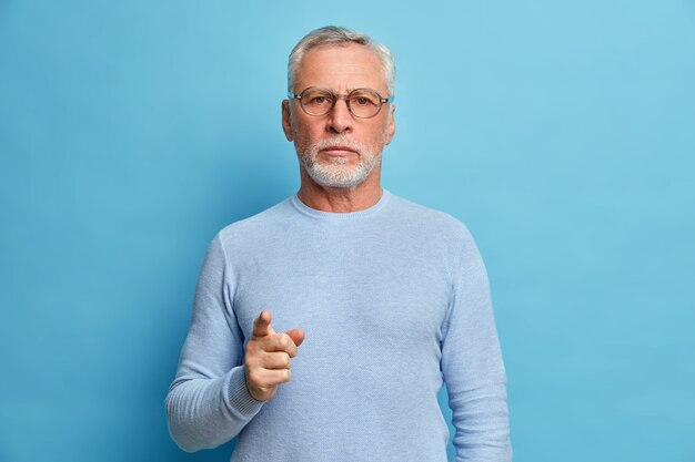 L'uomo barbuto maturo sicuro di sé indica davanti e seleziona che guardi seriamente la telecamera sceglie qualcuno nella sua squadra che indossa un maglione casual isolato sopra il muro blu