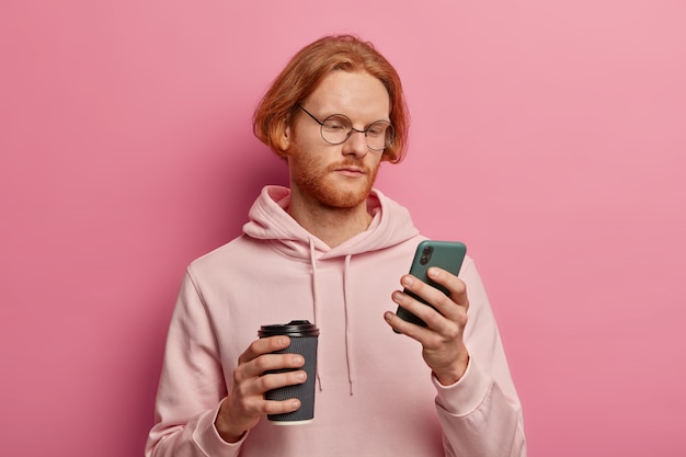 L'uomo barbuto serio utilizza il moderno telefono cellulare per la comunicazione online, controlla le e-mail, si concentra sullo schermo, beve caffè da asporto, indossa occhiali ottici e felpa con cappuccio, isolato sul muro rosa