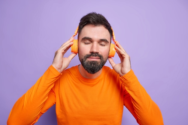 L'uomo barbuto soddisfatto riposa con la musica indossa le cuffie stereo sulle orecchie ascolta la playlist preferita tiene gli occhi chiusi indossa un maglione arancione brillante