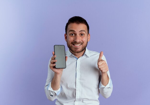 L'uomo bello emozionante tiene i pollici del telefono in alto isolati sulla parete viola