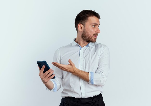 L'uomo bello scontento tiene e indica il telefono guardando il lato isolato sul muro bianco