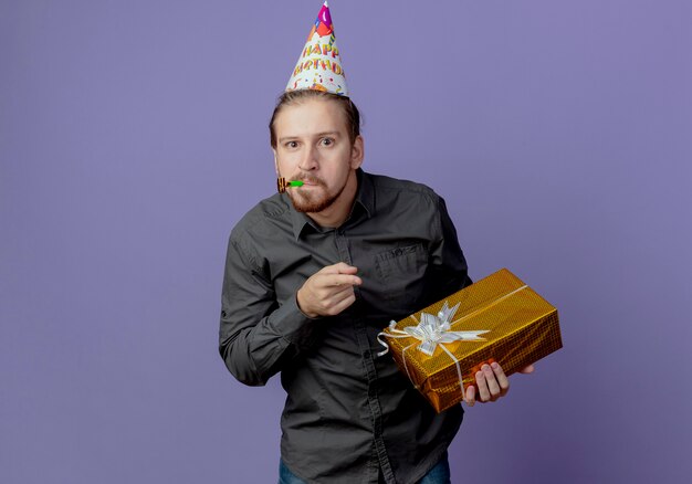 L'uomo bello sorpreso in berretto di compleanno tiene e indica il fischio di salto del contenitore di regalo isolato sulla parete viola