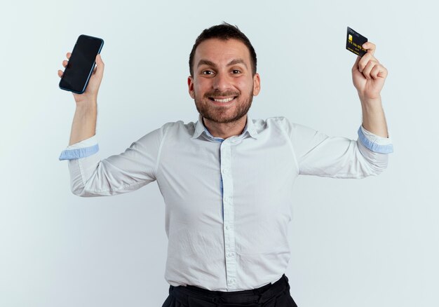 L'uomo bello sorridente alza le mani che tengono il telefono e la carta di credito isolata sulla parete bianca