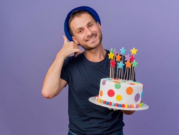 L'uomo bello sorridente che porta i gesti del cappello blu mi chiama segno e tiene la torta di compleanno isolata sulla parete viola con lo spazio della copia