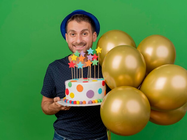 L'uomo bello sorridente che porta il cappello blu del partito tiene i palloni dell'elio e la torta di compleanno isolata sulla parete verde con lo spazio della copia