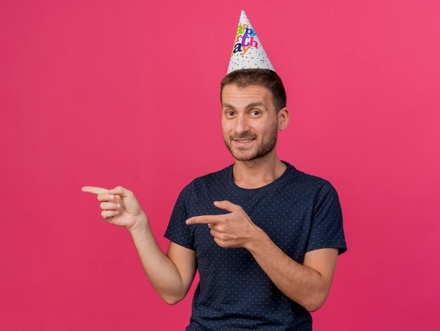 L'uomo bello sorridente che porta il cappuccio di compleanno punta a lato con due mani isolate sulla parete rosa con lo spazio della copia