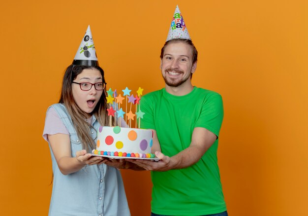 L'uomo bello sorridente in cappello del partito tiene la torta di compleanno e il cappello da portare della festa della ragazza sorpresa tiene e guarda la torta isolata