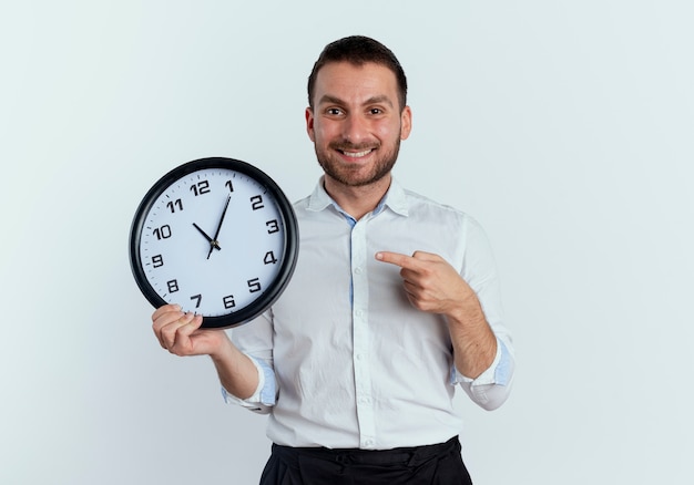 L'uomo bello sorridente tiene e indica l'orologio isolato sulla parete bianca