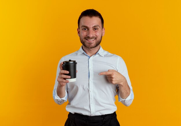 L'uomo bello sorridente tiene e indica la tazza di caffè isolata sulla parete arancione