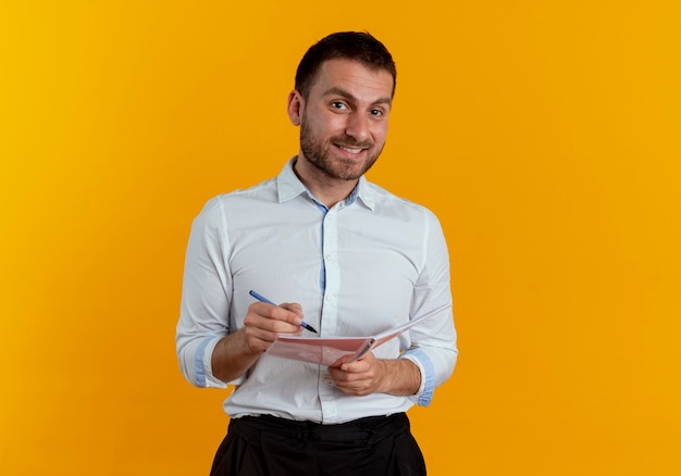 L'uomo bello sorridente tiene la penna ed il taccuino che sembrano isolati sulla parete arancione