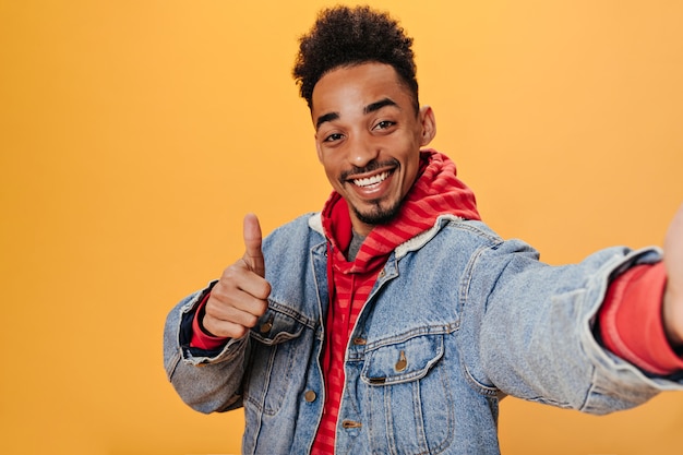 L'uomo castana in felpa con cappuccio e giacca di jeans mostra il pollice in su e fa selfie