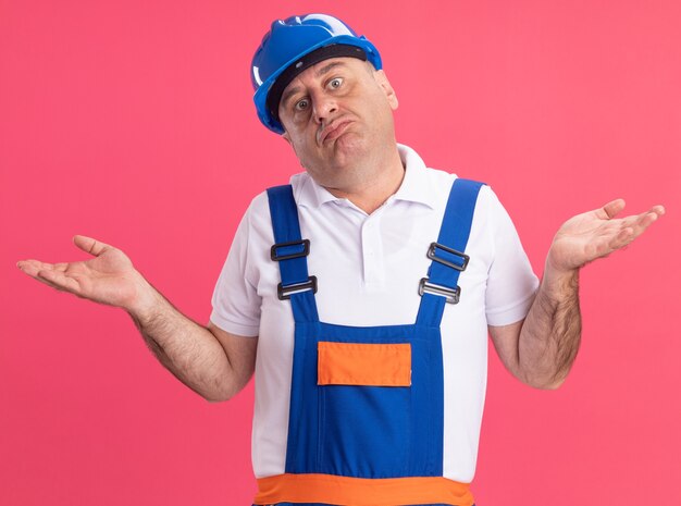 L'uomo caucasico adulto confuso del costruttore in uniforme tiene le mani aperte sul rosa