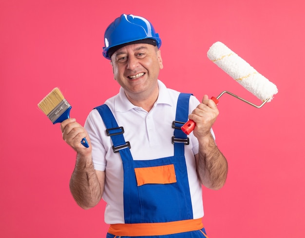 L'uomo caucasico adulto sorridente del costruttore in uniforme tiene il pennello e la spazzola del rullo sul colore rosa