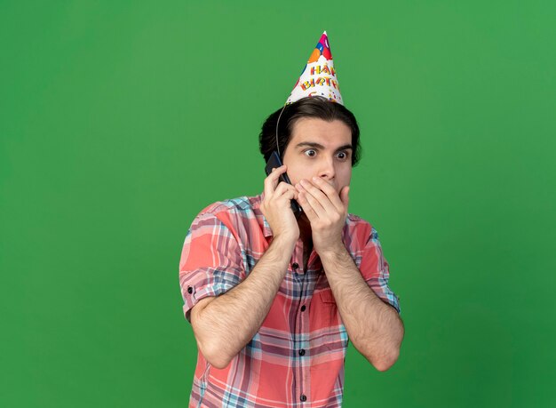 L'uomo caucasico bello sorpreso che indossa il berretto di compleanno mette la mano sulla bocca parlando al telefono