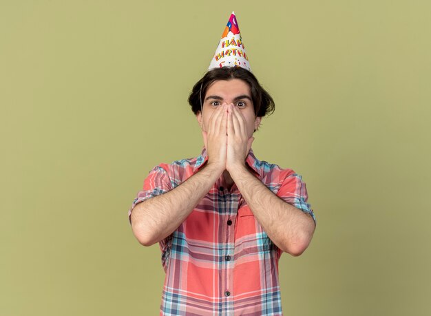 L'uomo caucasico bello sorpreso che indossa il berretto di compleanno mette le mani sulla bocca guardando la telecamera