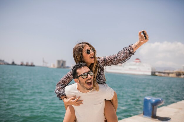 L&#39;uomo che dà alla sua ragazza sulle spalle ride prendendo selfie sul cellulare vicino al mare