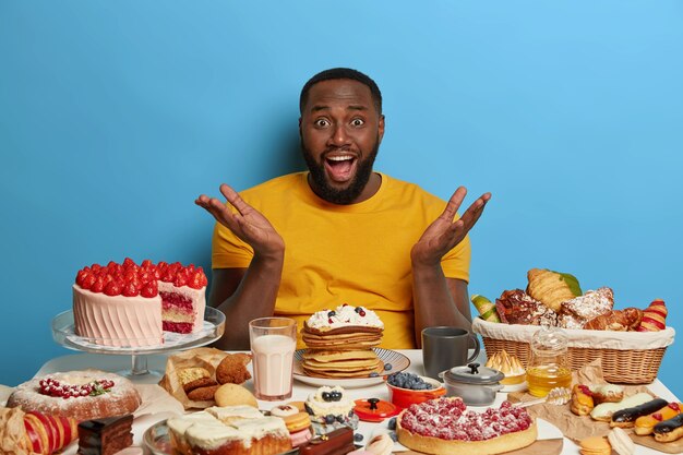 L'uomo con la barba addicted in sovrappeso ha un'espressione perplessa, allarga le palme, non vede l'ora di mangiare dolci, circondato da gustose torte, biscotti e latte