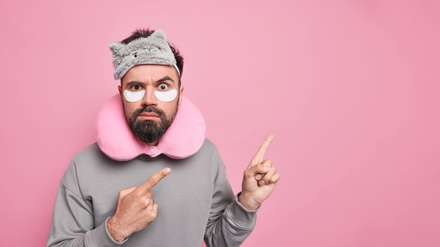 l'uomo con la barba folta punta in uno spazio vuoto applica cerotti sotto gli occhi per ridurre le rughe indossa un cuscino sul collo bendato