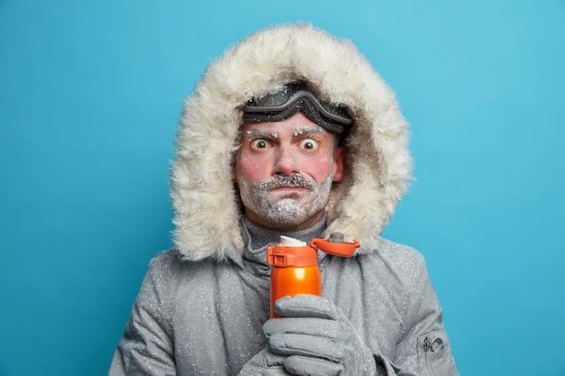 L'uomo congelato perplesso in abiti invernali cerca di scaldarsi con una bevanda calda ha la faccia rossa e l'orso coperto di bufera di neve trascorre molto tempo all'aperto durante lo snowboard. Condizioni meteorologiche gelide