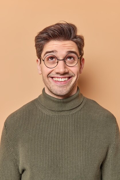 L'uomo curioso felice guarda con interesse sopra i sorrisi sente con gioia uno strano rumore al piano di sopra vestito posa casualmente sul muro beige dello studio. Soddisfatto ragazzo felice concentrato sorride a trentadue denti indossa gli occhiali