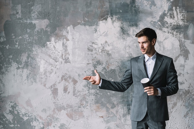 L&#39;uomo d&#39;affari che tiene la tazza di caffè asportabile che fa la pistola della mano gesture contro la parete del grunge