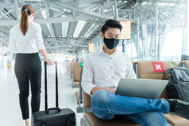 L'uomo d'affari di distanza sociale che indossa una maschera per il viso si siede a lavorare con il laptop mantenendo la distanza l'uno dall'altro per evitare l'infezione da covid19 durante la pandemia La croce rossa del sedile vuoto della sedia mostra una nuova normalità