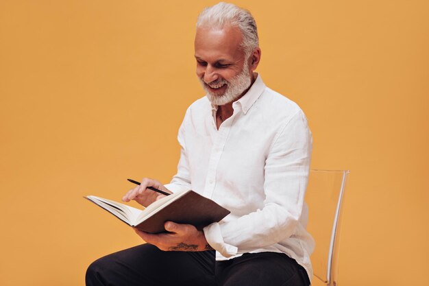 L'uomo dai capelli bianchi in camicia pone con il taccuino su sfondo arancione Affascinante adulto in abito stile ufficio guarda il blocco note e sorride