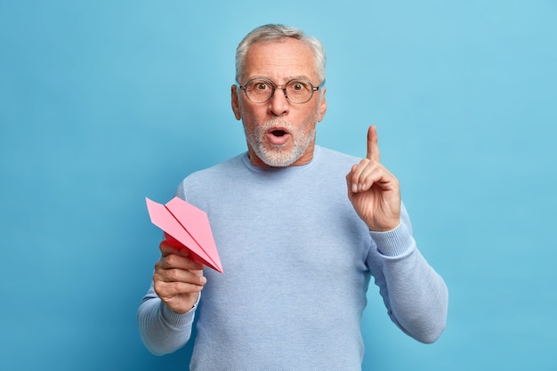 L'uomo dai capelli grigi maturo barbuto sorpreso e sbalordito punta il dito indice in alto ha un'idea eccellente tiene l'aereo di carta tiene la bocca aperta indossa occhiali e pose del ponticello contro il muro blu dello studio