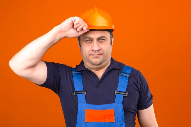 L'uomo del costruttore che indossa l'uniforme della costruzione e il casco di sicurezza che tengono il suo casco gradiscono il saluto con il fronte serio sopra la parete arancio isolata