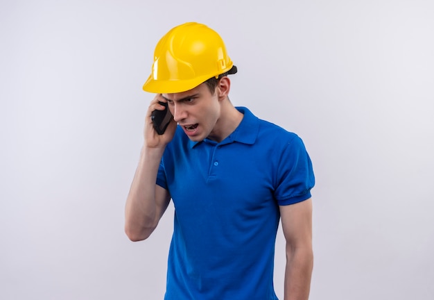 L'uomo del giovane costruttore che indossa l'uniforme della costruzione e il casco di sicurezza parla con rabbia al telefono