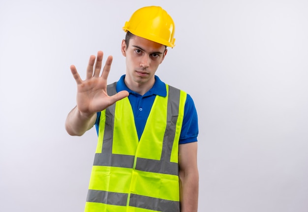 L'uomo del giovane costruttore che indossa l'uniforme della costruzione e il casco di sicurezza si ferma seriamente con la mano