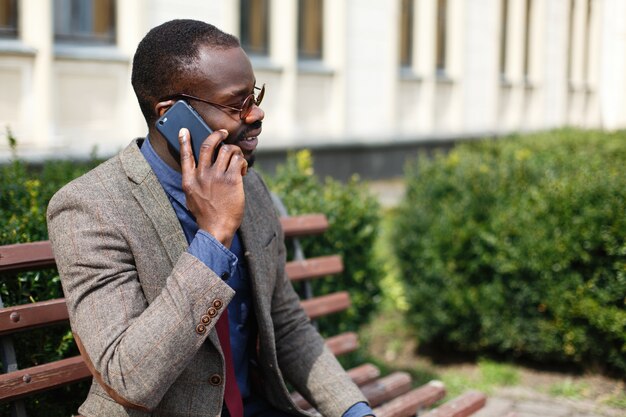 L&#39;uomo di affari dell&#39;afroamericano comunica sullo smartphone che si siede sul banco all&#39;esterno