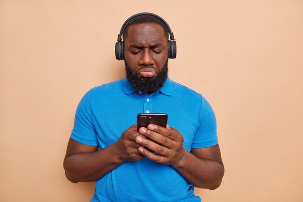 L'uomo dispiaciuto con la barba folta tipi di messaggi di testo tramite smartphone infelice di leggere commenti negativi sotto il post ascolta musica tramite le cuffie indossa una maglietta blu pone un muro marrone al coperto