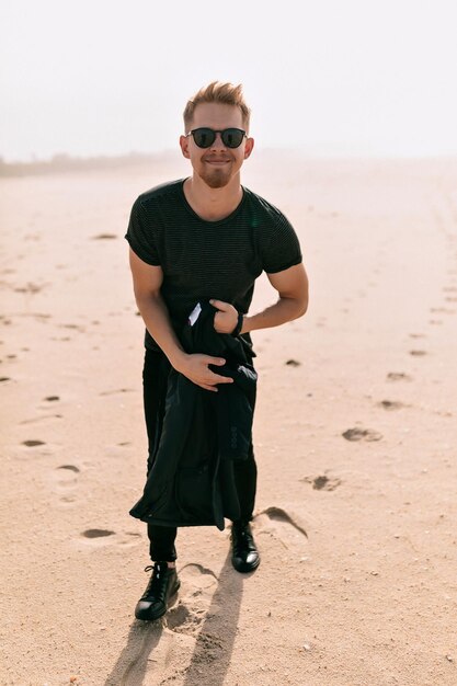 L'uomo eccitato divertente in maglietta nera sta tenendo la giacca nelle mani è in posa davanti alla macchina fotografica con un sorriso felice mentre cammina sulla spiaggia vicino all'oceano