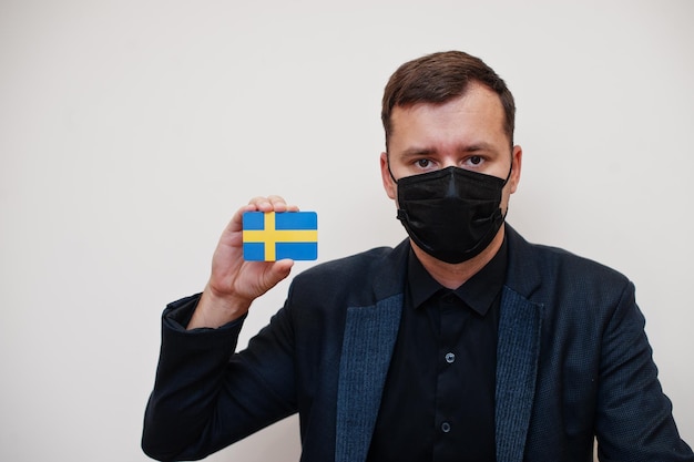 L'uomo europeo indossa la maschera facciale nera e protegge la carta della bandiera svedese isolata su sfondo bianco Europa coronavirus Covid concetto di paese