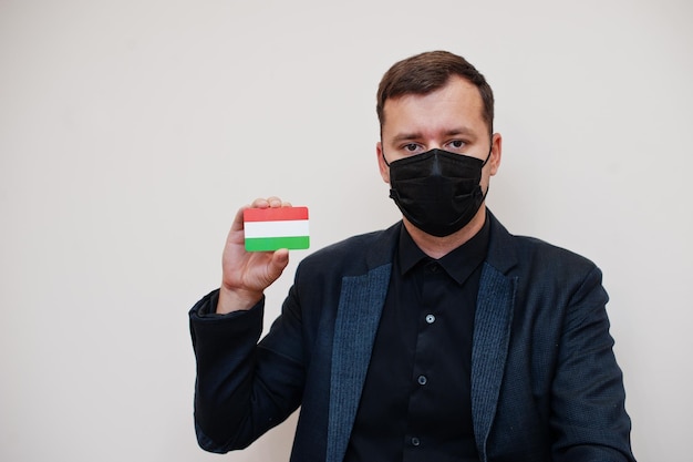 L'uomo europeo indossa una maschera nera formale e protegge il viso, tiene la carta della bandiera dell'Ungheria isolata su sfondo bianco Europa coronavirus Covid concetto di paese