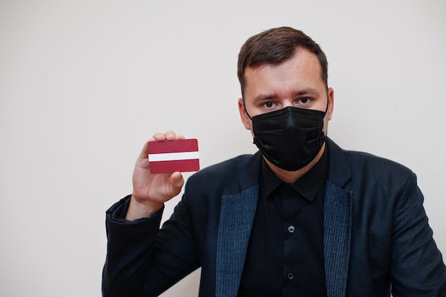 L'uomo europeo indossa una maschera nera formale e protegge il viso, tiene la carta della bandiera della Lettonia isolata su sfondo bianco Europa coronavirus Covid concetto di paese