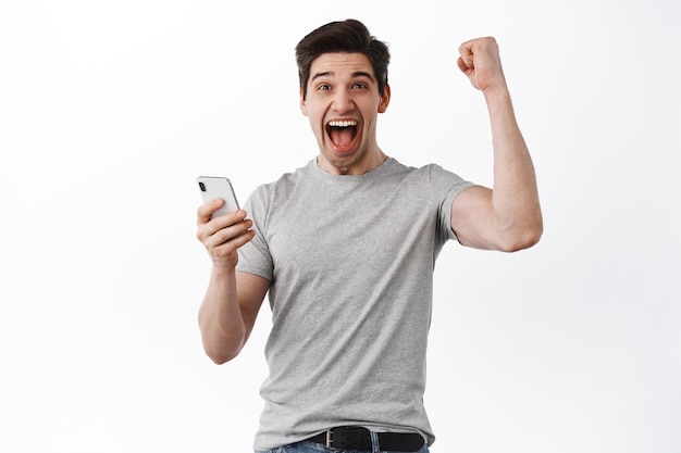 L'uomo felice tiene lo smartphone e celebra, vince il premio online, grida sì e si rallegra, trionfa dal successo, sembra soddisfatto, sfondo bianco