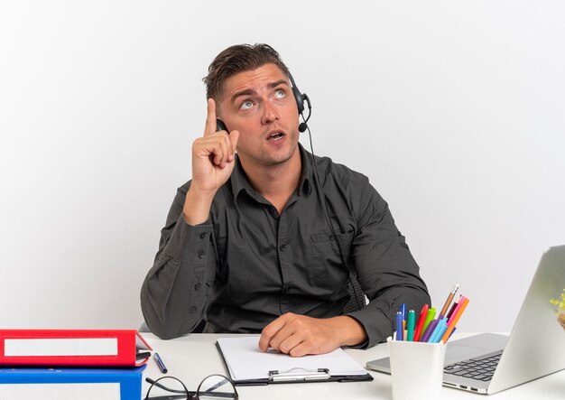 L'uomo giovane sorpreso dell'operaio di ufficio biondo sulle cuffie si siede alla scrivania con gli strumenti dell'ufficio utilizzando gli sguardi del computer portatile e indica in alto isolato su priorità bassa bianca con lo spazio della copia