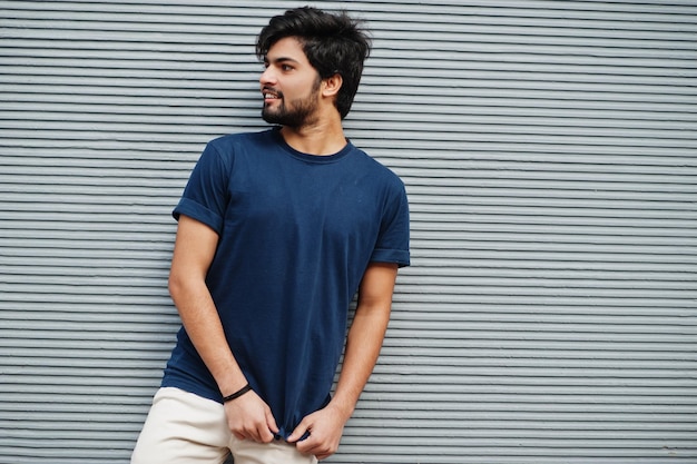 L'uomo indiano casual alla moda indossa una maglietta blu in posa contro il muro grigio